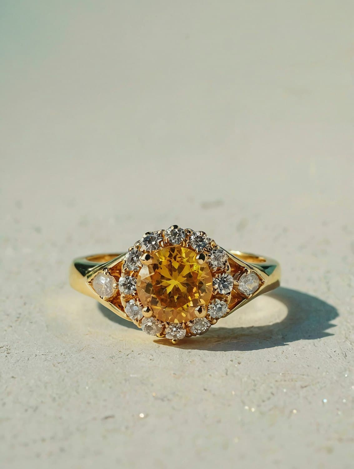 A gold ring with a round yellow center stone ringed by small white stones, casting a soft shadow on pale stone.