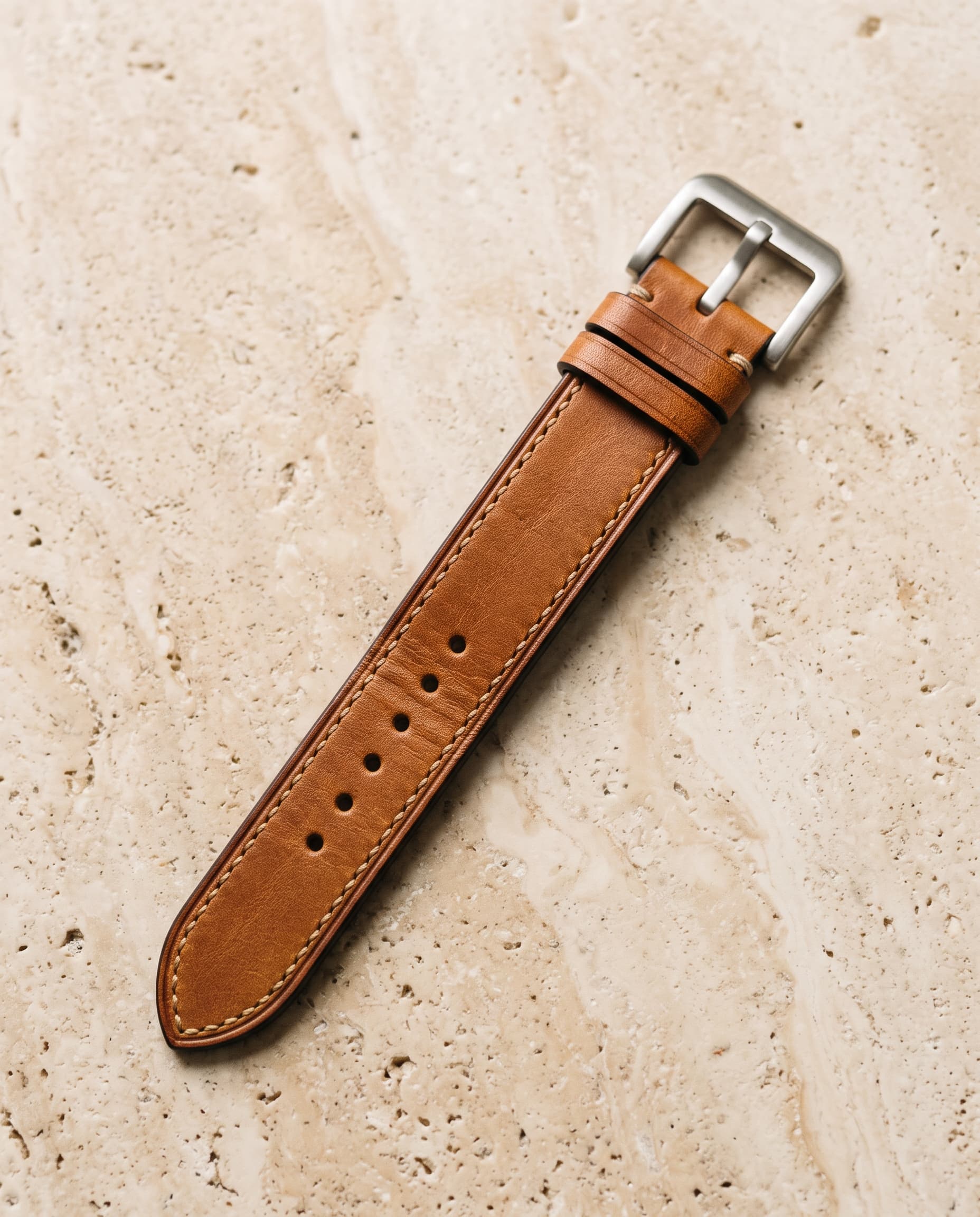 A tan calf leather watch strap with stitched edges and a brushed steel buckle, laid flat on pale stone.