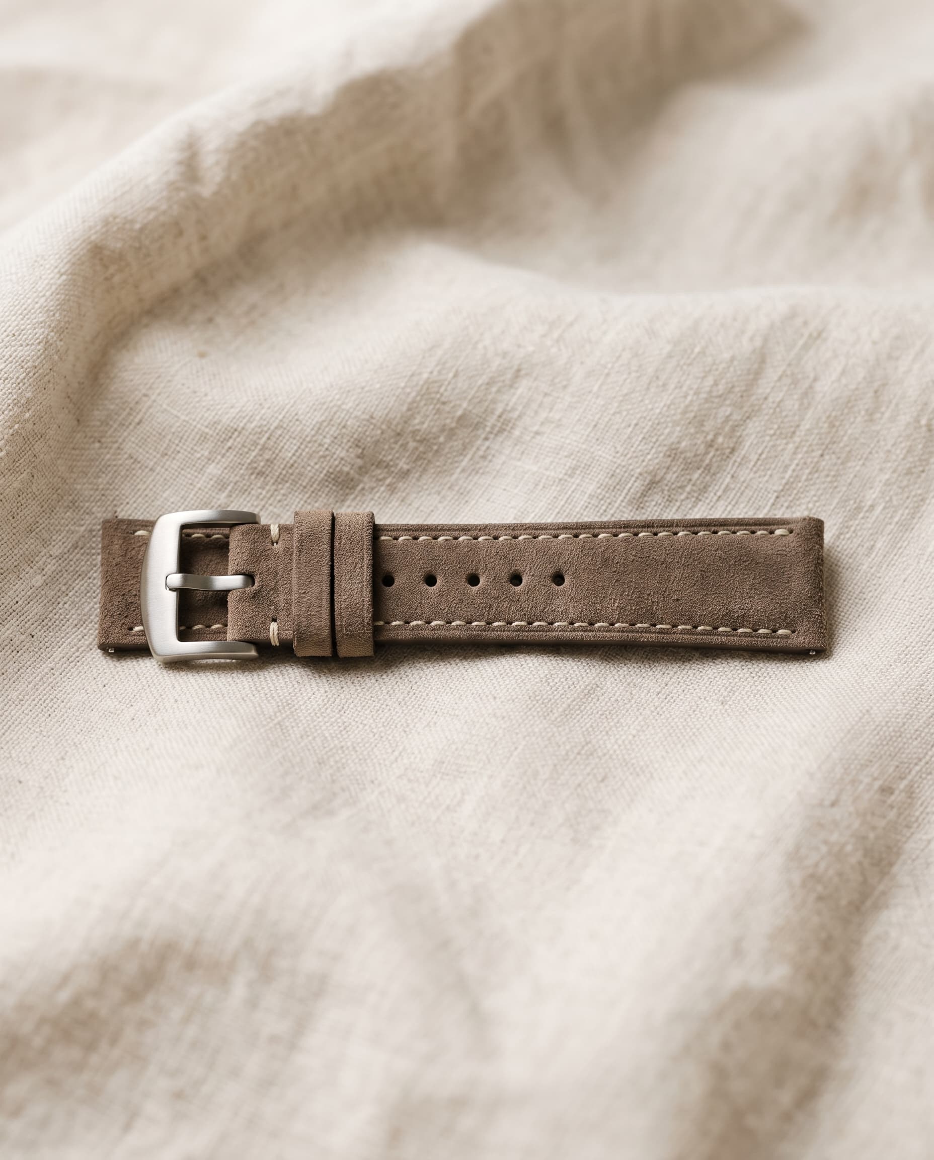 A taupe suede watch strap with ivory stitching and a brushed steel buckle, resting on cream linen.