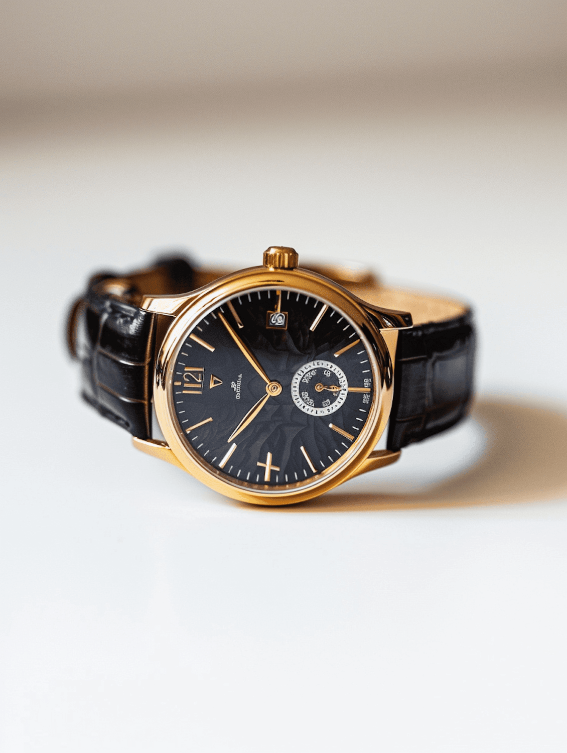 A gold watch with a black textured dial, a small seconds sub-dial and a black leather strap, resting on a white surface.