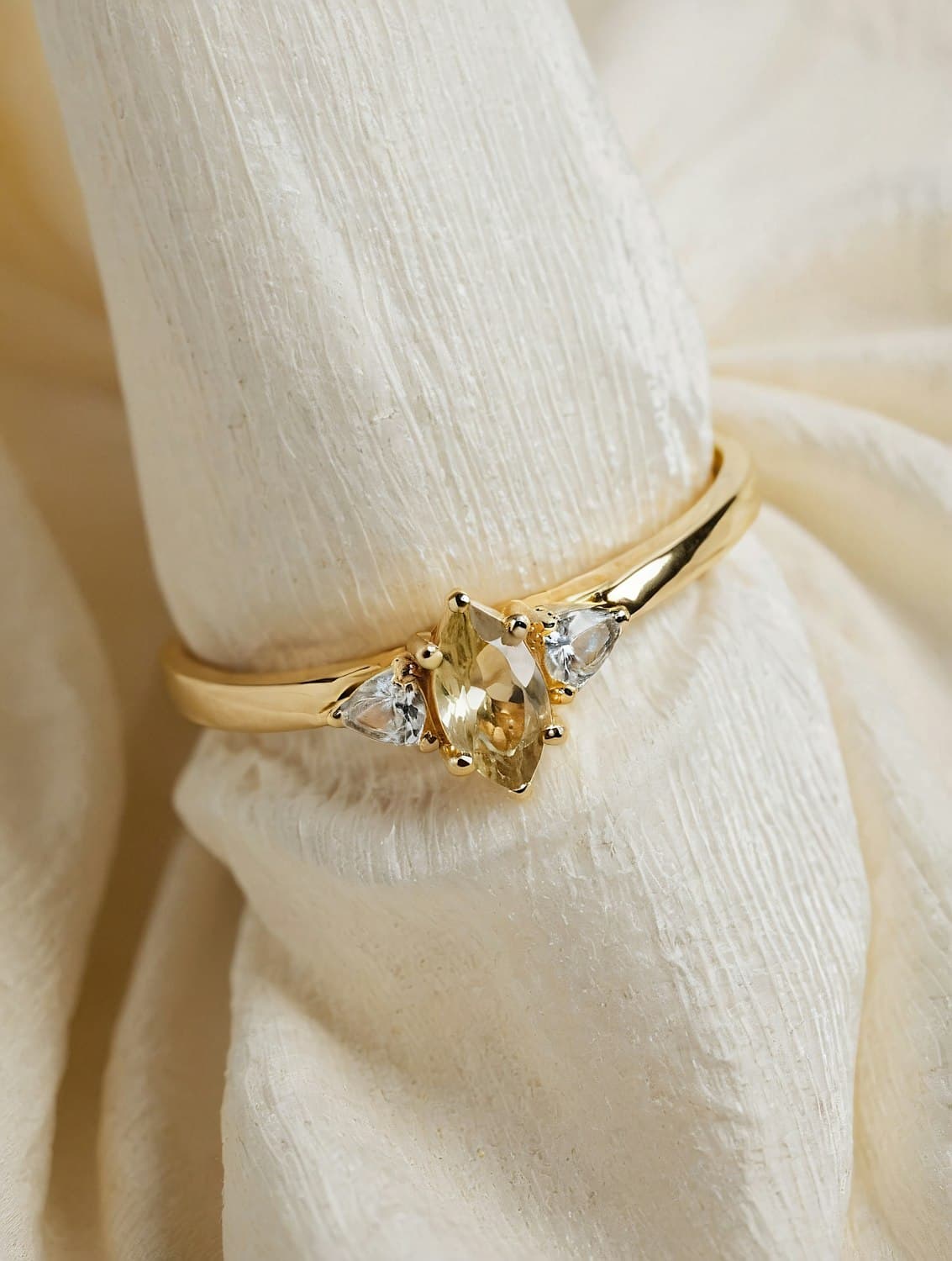 Close view of a gold ring whose claws hold a pale marquise stone between two white trillion-cut stones.